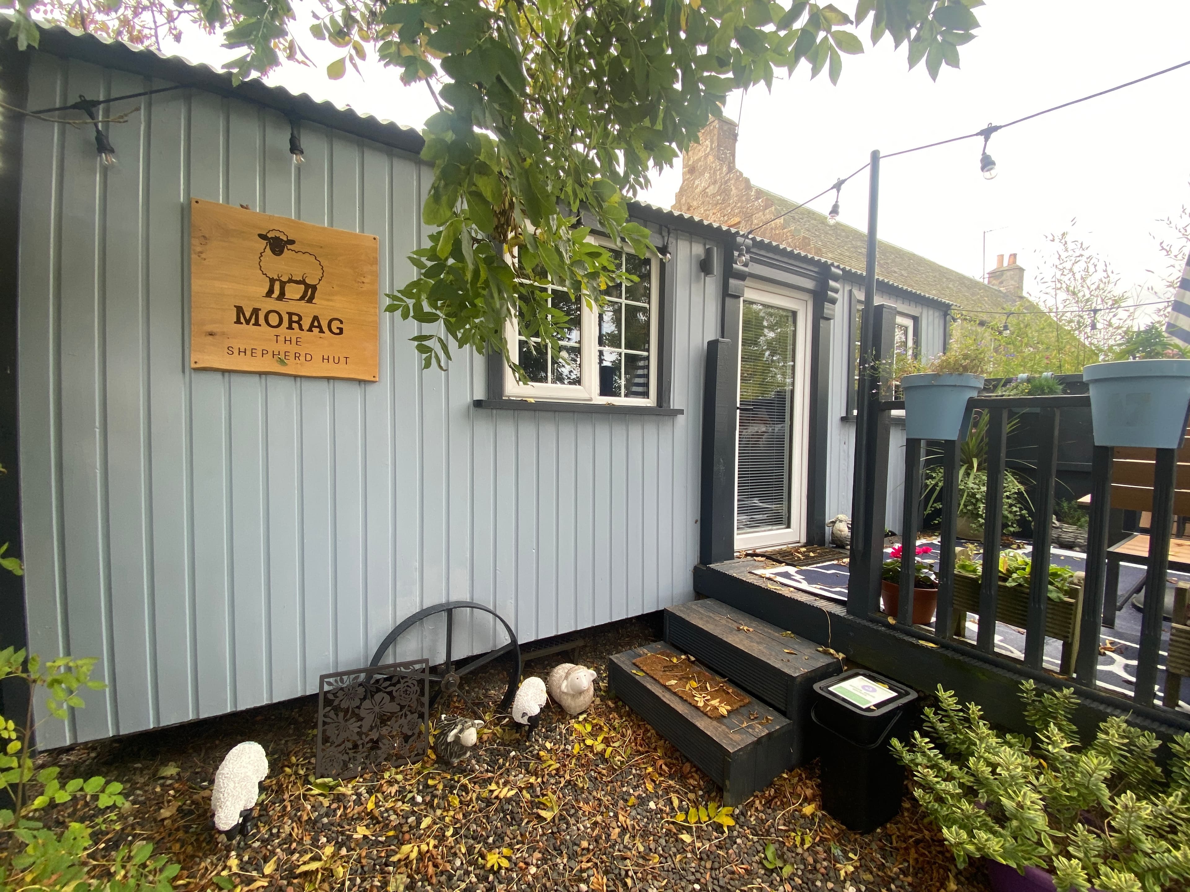 Morag the shepherd hut in a St Andrews garden, with muted blue and grey timber cladding, white window frames and charcoal trim, steps and decking.