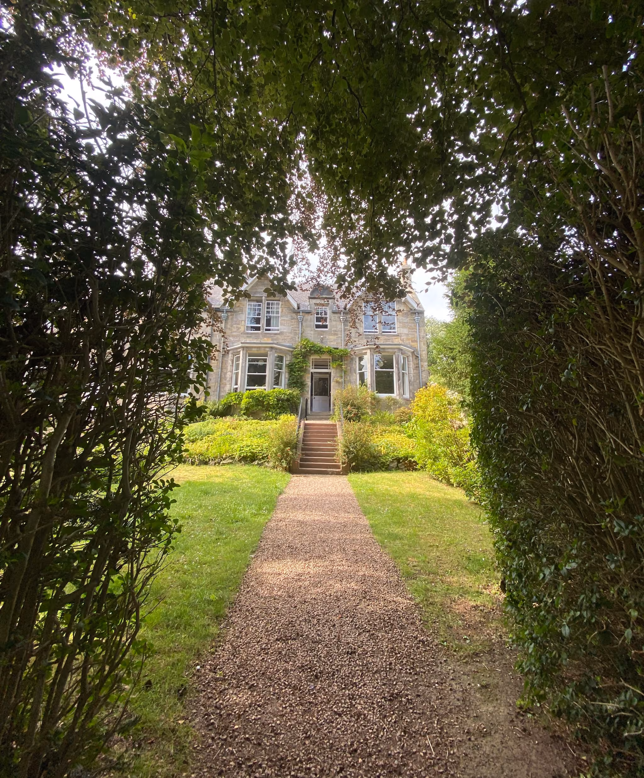 Tree lined gravel path leading through a green garden to Emilie's traditional stone villa in St Andrews.